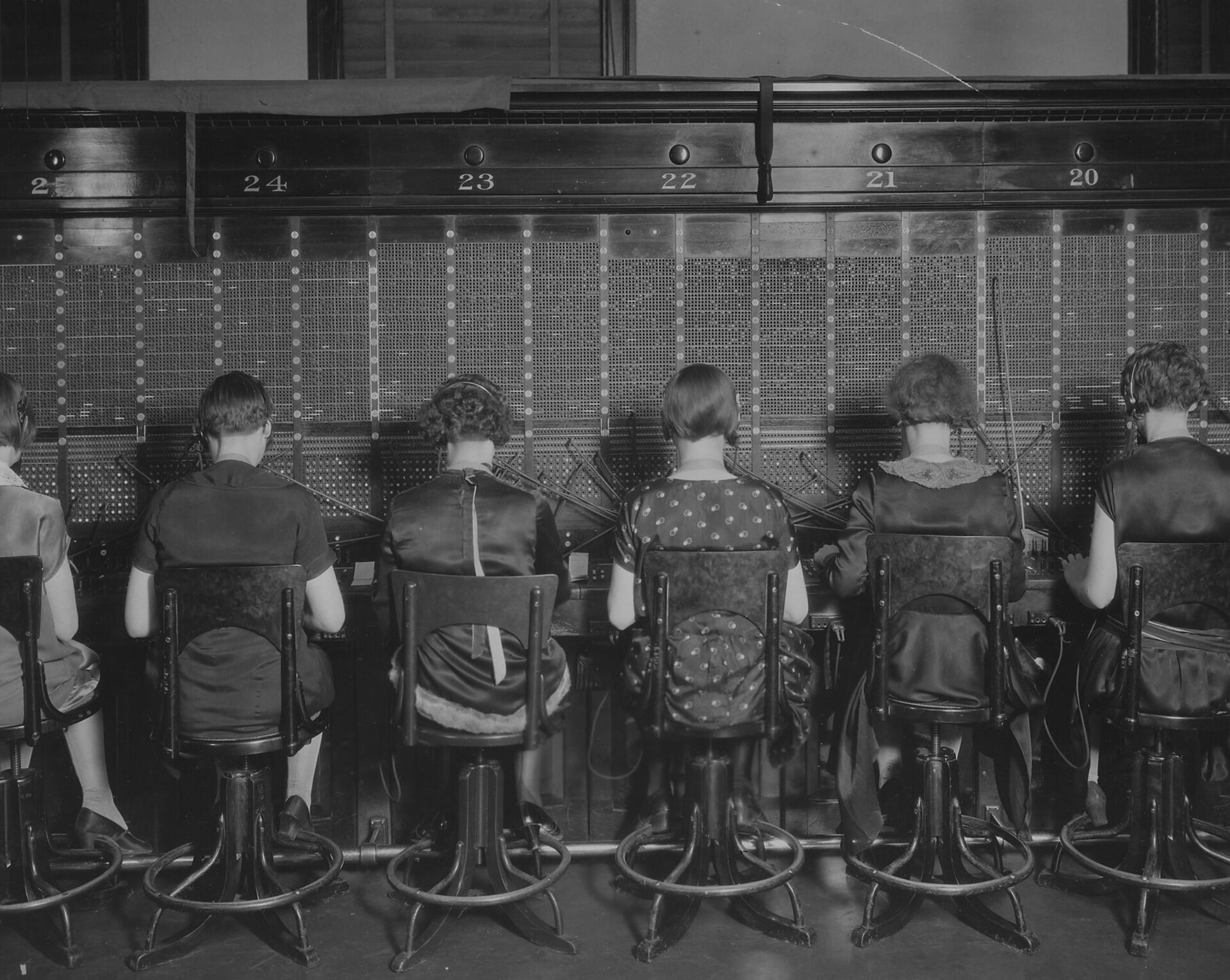 Six telephone operators seen from behind, seated in a row at a large switchboard, headsets on, plugging in calls